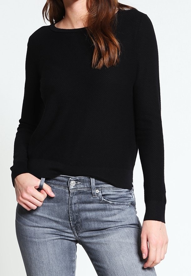 Kobieta w czarnym swetrze z długim rękawem o teksturowanej fakturze i wyblakłych szarych dżinsach, z jedną ręką trzymającą pętlę od pasa.