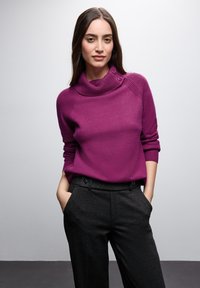 Fioletowy sweter z wysokim kołnierzem, z teksturowanymi akcentami na ramionach i detalami z guzikami, zestawiony z ciemnymi, dopasowanymi spodniami.