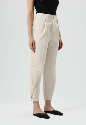 Beige hoge taille broek met plooien, taps toelopende pijpen en zijzakken. Heeft een knopdetail bij de enkel. Draagt zwarte instapschoenen.