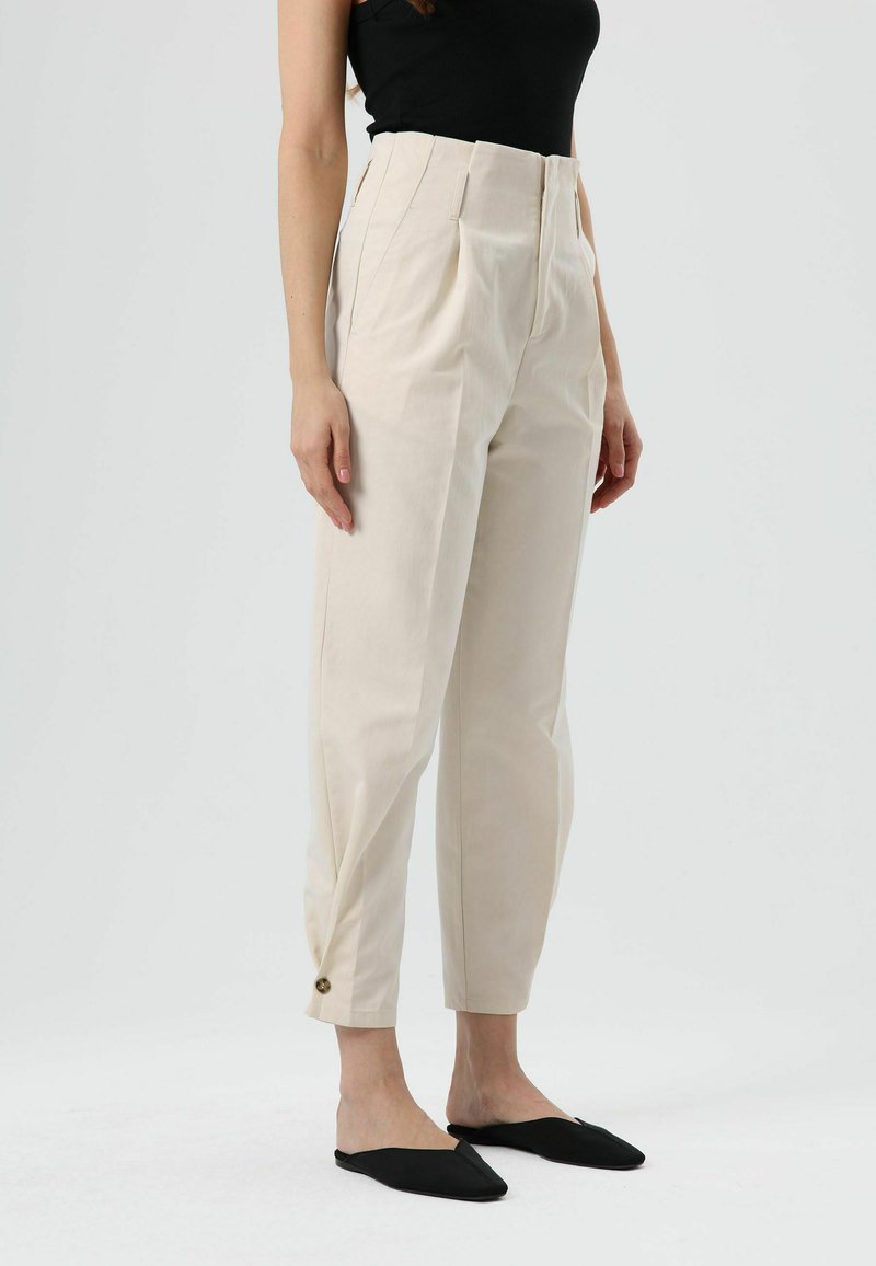 Beige, hoch taillierte Hose mit Bügelfalten, schmal zulaufenden Beinen und seitlichen Taschen. Mit einer Knopfverzierung am Knöchel. Kombiniert mit schwarzen Slippern.