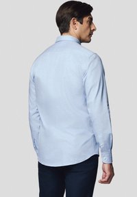 Jasnoniebieska, długosłębowa koszula z kołnierzykiem zapinanym na guziki i zaokrąglonym dołem. Materiał wydaje się gładki, z subtelną teksturą. Noszona z ciemnymi spodniami.