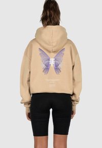 Beige Kapuzenpullover mit einem großen lila Schmetterlingsmotiv auf dem Rücken. Verfügt über eine entspannte Passform und eine Kordelzugkapuze. Schwarze Radlerhosen werden darunter getragen.