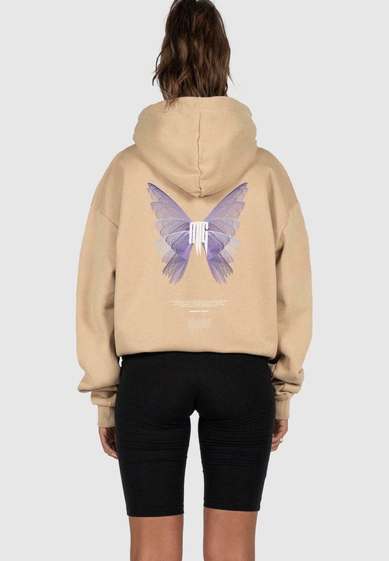 Beige Kapuzenpullover mit einem großen lila Schmetterlingsmotiv auf dem Rücken. Verfügt über eine entspannte Passform und eine Kordelzugkapuze. Schwarze Radlerhosen werden darunter getragen.