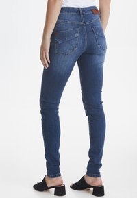 Jeans de denim azul delgado con una cintura alta, que cuentan con bolsillos traseros y una etiqueta de cuero marrón. Textura suave con un ligero estiramiento.