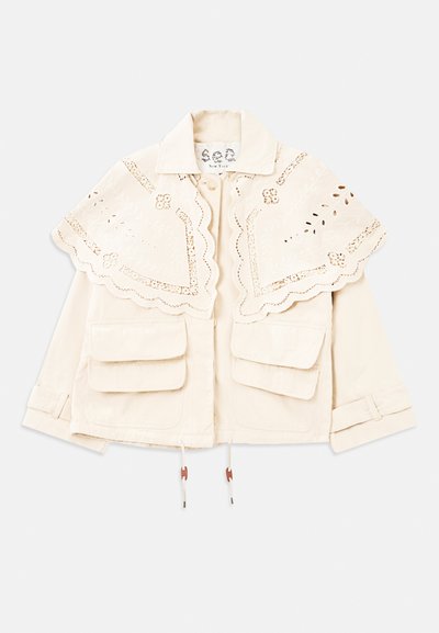 Kreminė striukė su išpjaustytu, siuvinėtų apykakle, dviem priekinėmis kišenėmis su sklende, mygtukų uždarymu ir reguliuojamais rankogaliais.