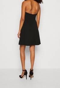 Czarna sukienka bez ramiączek z dopasowanym gorsetem i rozkloszowaną spódnicą. Gładki materiał z subtelnym połyskiem. Połączona z czarnymi szpilkami.