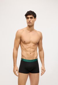 Boxer shorts de algodón negros con una cinturilla de logo verde, llevados por un modelo que se encuentra en un entorno neutro, mostrando un diseño ajustado.