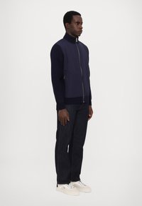 Navy zip-up jacket with ribbed knit collar and cuffs, paired with dark denim jeans and white sneakers. Smooth fabric with contrasting textures.