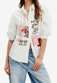 White cotton shirt featuring a patchwork design with stripes and character prints, including Mickey Mouse, paired with light blue jeans.