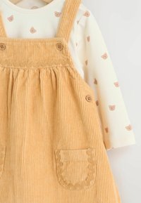 Wattierte Latzkleid aus weichem Gelb mit Rüschenspitze, zwei Taschen und Knopfverschlüssen, kombiniert mit einem cremefarbenen Langarmshirt mit Bärenmotiven.