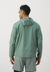Veste à capuche légère de couleur vert doux, avec une texture lisse, des manches longues et une coupe décontractée. Détails cousus le long des épaules.
