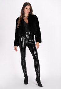 Schwarze Kunstfell-Cropped-Jacke über einem schwarzen, taillierten Bodysuit, kombiniert mit hochglänzenden, schwarzen Vinylleggings und schwarzen Ankle Boots.