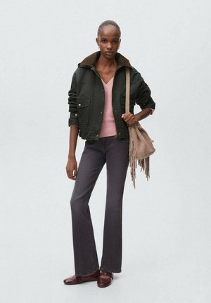 Donna che indossa una giacca verde scuro, un maglione rosa, pantaloni grigi svasati, scarpe color bordeaux e porta una borsa beige con frange a tracolla.