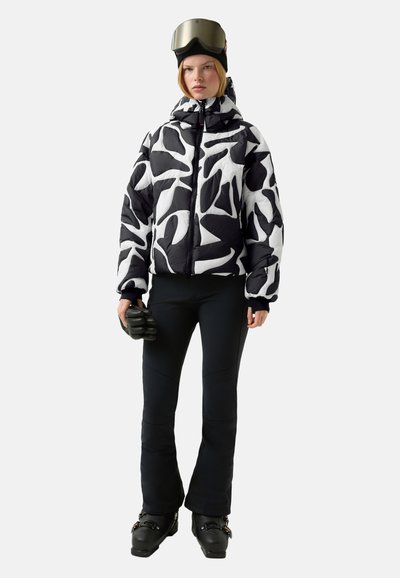 Pufová bunda s černobílým abstraktním vzorem, v kombinaci s černými kalhotami, rukavicemi a pletenou čepicí, se lyžařskou helmou a brýlemi.