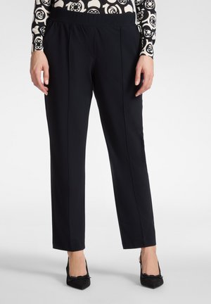 Donna che indossa pantaloni neri a gamba dritta con pieghe anteriori e tacchi neri a punta con dettaglio a fiocco, in piedi su sfondo bianco.