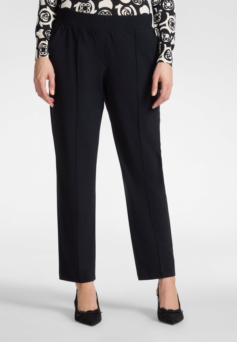 Donna che indossa pantaloni neri a gamba dritta con pieghe anteriori e tacchi neri a punta con dettaglio a fiocco, in piedi su sfondo bianco.