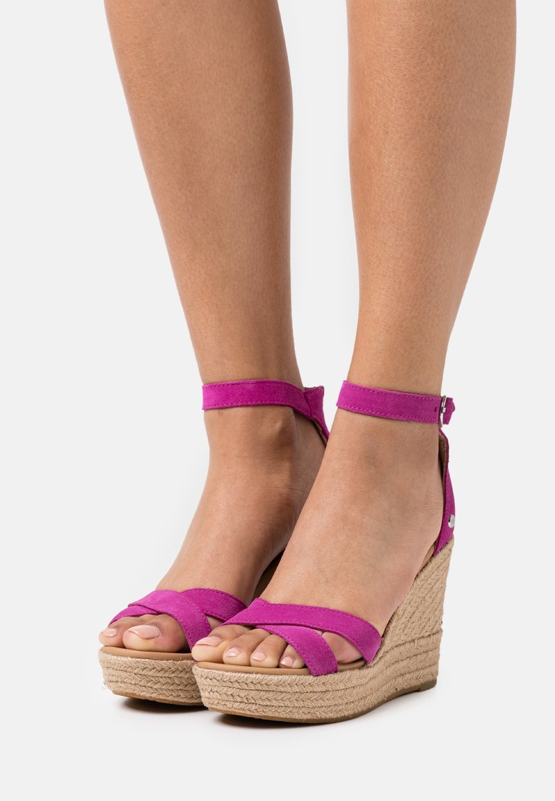 Klínové sandály s růžovým semišovým svrškem, křížovými popruhy, uzavřeným zadním dílem a tkaným jútovým podpatkem. Mají přezku na kotníku.