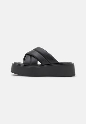 Sort platformsandal med brede krydsede polstrede stropper og tyk sål mod en ensfarvet lys baggrund.