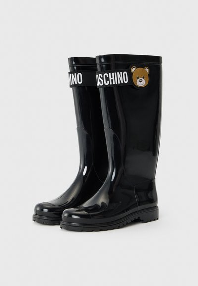 Stivali da pioggia neri in gomma con suola piatta, con una fascia bianca con la scritta "MOSCHINO" e un emblema di un orsetto marrone sul lato.