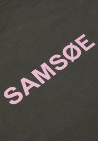 Mørkegrått stoff med en teksturert overflate. Har fet, lyse rosa tekst som staver "SAMØE" med en sirkulær aksent over bokstaven "O."