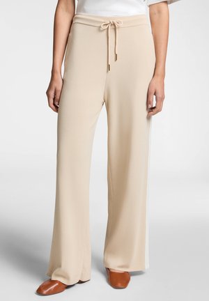Pantaloni palazzo beige realizzati in un tessuto morbido con vita regolabile e strisce bianche sui lati. Abbinati a scarpe basse marroni.