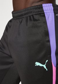 Čierne športové nohavice s hladkou textúrou, s fialovými a ružovými bočnými pruhmi a svetlo modrým logom Puma na ľavom boku.
