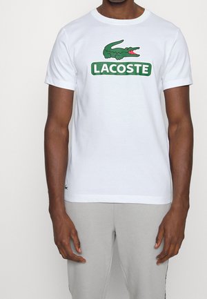 Mann trägt ein weißes kurzärmeliges T-Shirt mit grünem Krokodil-Logo und dem Schriftzug "LACOSTE" auf der Brust, kombiniert mit hellgrauer Hose.