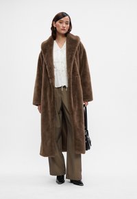 Casaco longo castanho e peludo com um colarinho largo, usado sobre uma blusa branca e calças largas bege, complementado por uma carteira preta.