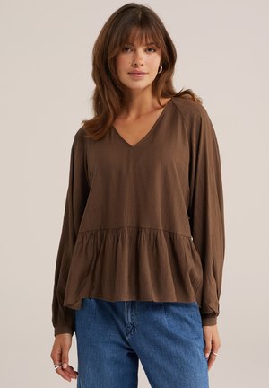 Kvinde iført en løs brun bluse med lange ærmer og V-hals og blå jeans mod en ensfarvet lys baggrund.