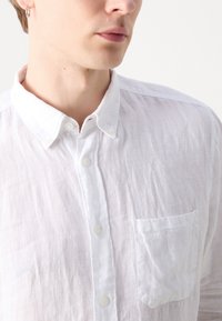 Weißes Leinenhemd mit Button-Down-Kragen, Brusttasche und strukturierter, leicht knitteriger Oberfläche. Verfügt über weiße Knöpfe und einen lockeren Schnitt.