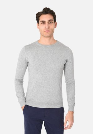 Uomo che indossa un maglione aderente a maniche lunghe di colore grigio chiaro e pantaloni blu scuro, in piedi con una mano in tasca su sfondo bianco.