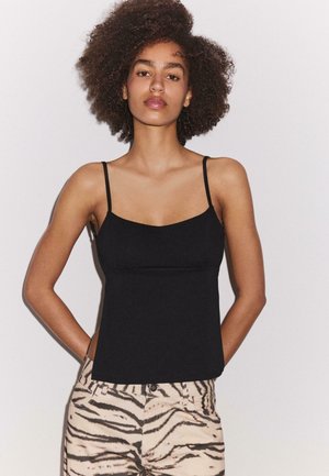 Femme aux cheveux bouclés portant un top noir à fines bretelles et un pantalon beige imprimé zèbre, debout les mains dans le dos.