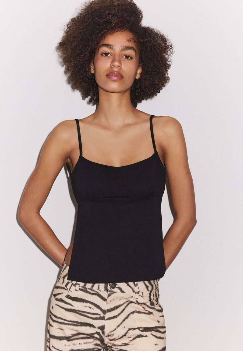 Femme aux cheveux bouclés portant un top noir à fines bretelles et un pantalon beige imprimé zèbre, debout les mains dans le dos.