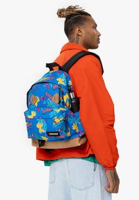 Mochila azul con patrones tropicales, correas de tela y un bolsillo frontal. Presenta un acento marrón en la base y una insignia con el logo.