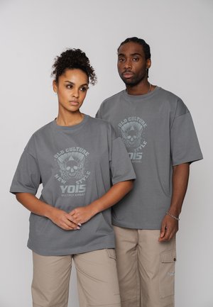 Two adults wearing matching gray graphic t-shirts and beige cargo pants, standing against a plain light gray background.