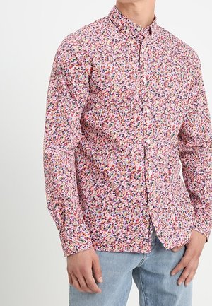 Man wearing a long-sleeve, buttoned shirt with a multicolored small floral pattern and light blue jeans, standing against a white background.