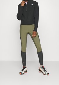 Schwarze Zip-Up-Sweatjacke mit gerippten Bündchen, kombiniert mit olivgrünen Leggings mit schwarzen Akzenten und einer enganliegenden Passform. Schuhe mit weißen, schwarzen und orangen Details.