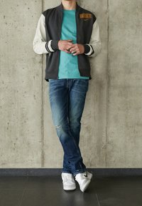 Grå zip-up jacka med vita ärmar, turkos t-shirt, slitna blå jeans och vita sneakers med svarta accenter, mot en betongvägg.