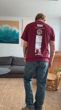 Homem com uma t-shirt burgundy da Levi's e jeans azuis está em uma sala de estar com um sofá cinza, mesa de vidro, cadeira de vime e arte de parede abstrata.