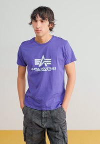 Purpura kokvilnas T-kreklis ar baltu logo, uz kura rakstīts "ALPHA INDUSTRIES" un trīs svītras zemāk, kombinēts ar pelēkajām cargo šortiem.