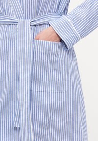 Ljusblå och vit randig morgonrock med bälte i midjan, långa ärmar och en ficka på vänster sida. Tillverkad av bomull med en mjuk textur.