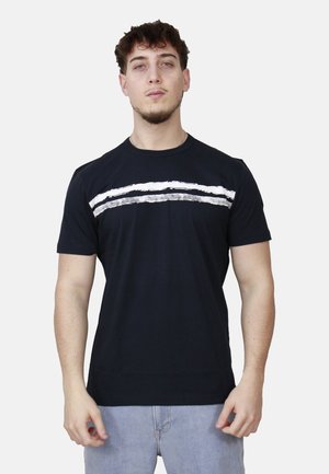 Giovane uomo che indossa una t-shirt nera a maniche corte con due strisce orizzontali bianche a pennellate sul petto, abbinata a jeans azzurri.