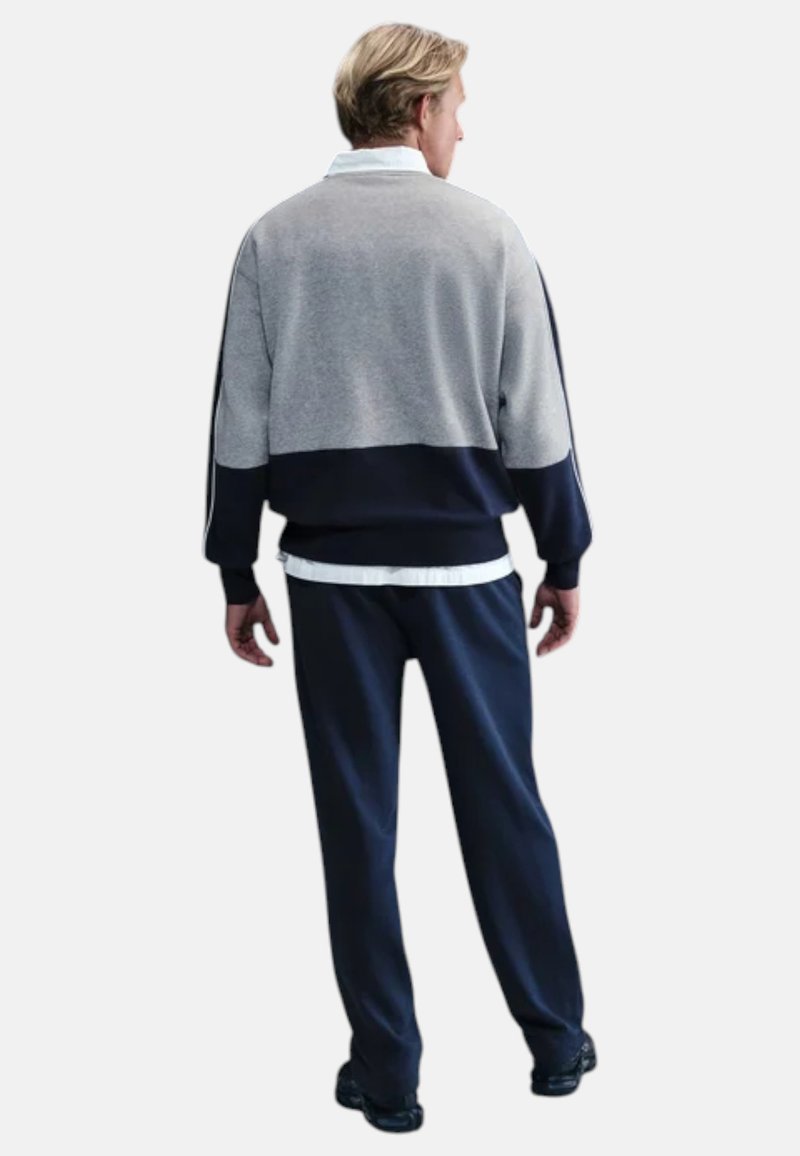 Uomo che indossa una felpa a blocchi di colore grigio e blu navy, pantaloni blu navy, camicia bianca sotto e scarpe nere, in piedi di spalle.