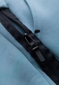 Jasnoniebieski materiał z kapturem, z frontowym zamkiem błyskawicznym. Czarny zamek i uchwyt z brandingiem "OMBRE". Widoczny teksturowany materiał.