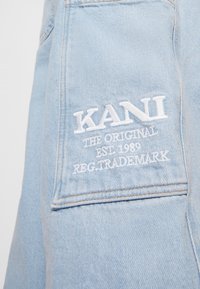 Svetle modrá džínsovina s přišitým kapsou, na které je bíle vyšitý text: KANI THE ORIGINAL EST. 1989 REG. TRADEMARK.