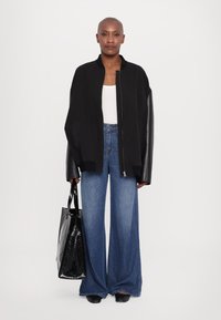 Black bomber jacket with leather sleeves, white tank top, wide-leg blue jeans, holding a shiny black tote bag, standing against a white background.