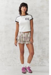 Weißes, verkürztes T-Shirt mit schwarzem Rand und Druck "01", kombiniert mit karierter Rüschenshorts in Pink, Grün und Weiß. Getragen mit rosa Turnschuhen.