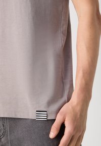 T-shirt gris avec une texture lisse, présentant une étiquette rayée noir et blanc sur l'ourlet. Gros plan sur le côté avec une main qui le tient.