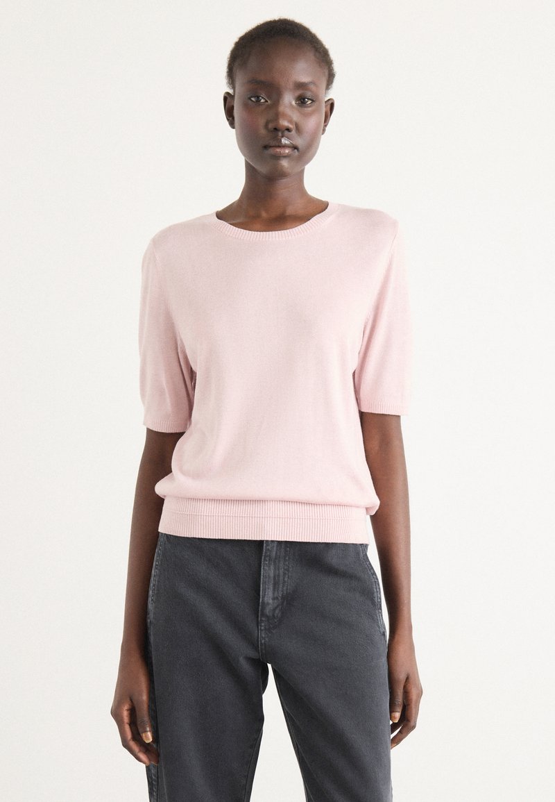 Short-sleeve pink knit sweater with a ribbed crew neckline and hem. Worn with dark grey jeans, featuring a relaxed fit and smooth texture.