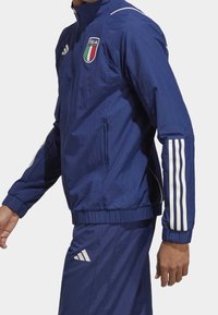 Marineblå vindjakke med fuld lynlås og høj krave; har hvide Adidas-striber på ærmerne og det italienske nationale emblem på brystet.